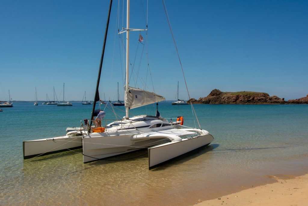 Un catamaran à voile se repose sur une plage de sable fin aux eaux bleues et limpides, sous un ciel ensoleillé.